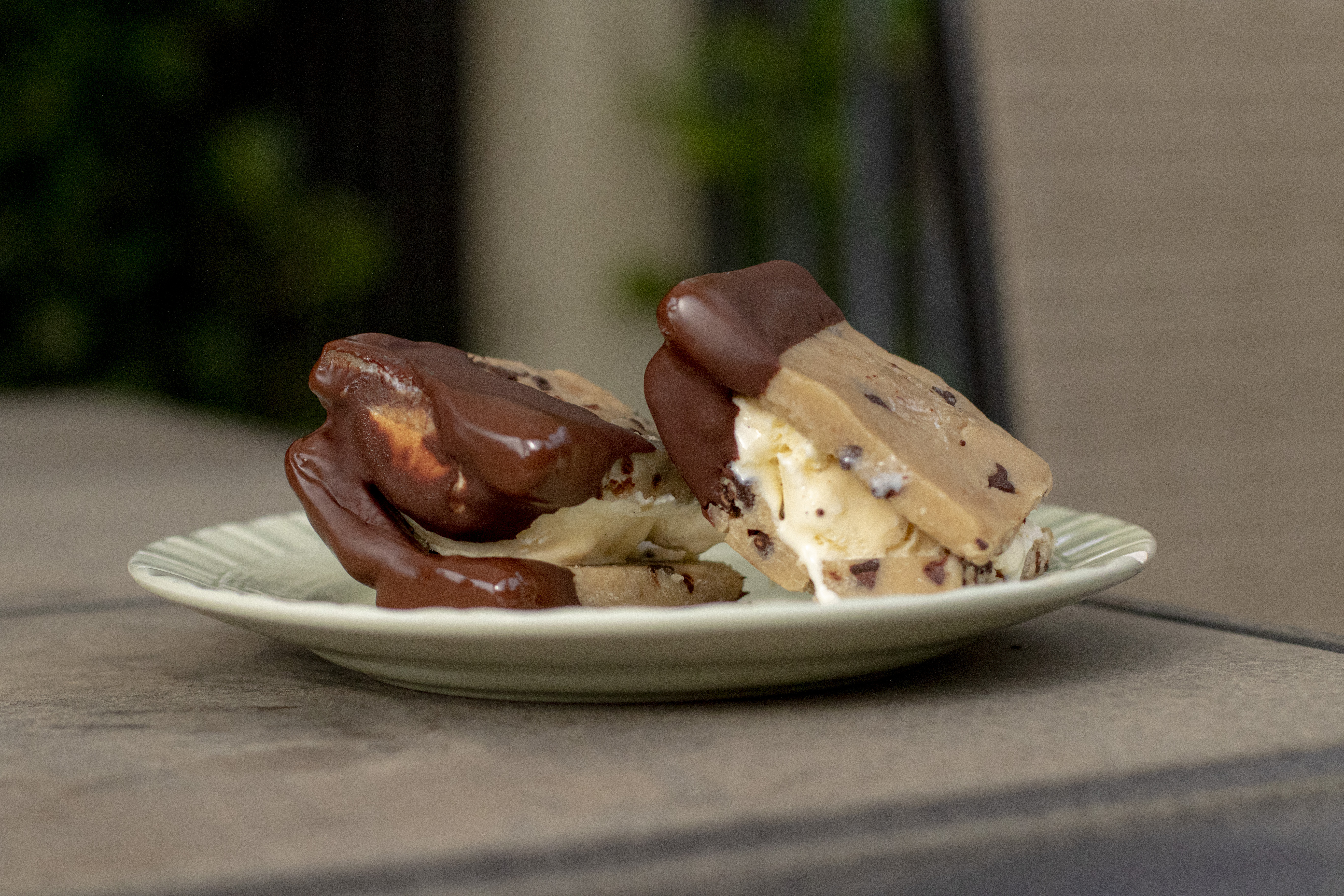 cookie dough ice cream sandwiches coated in chocolate on a green plate sitting on a patio table outside.