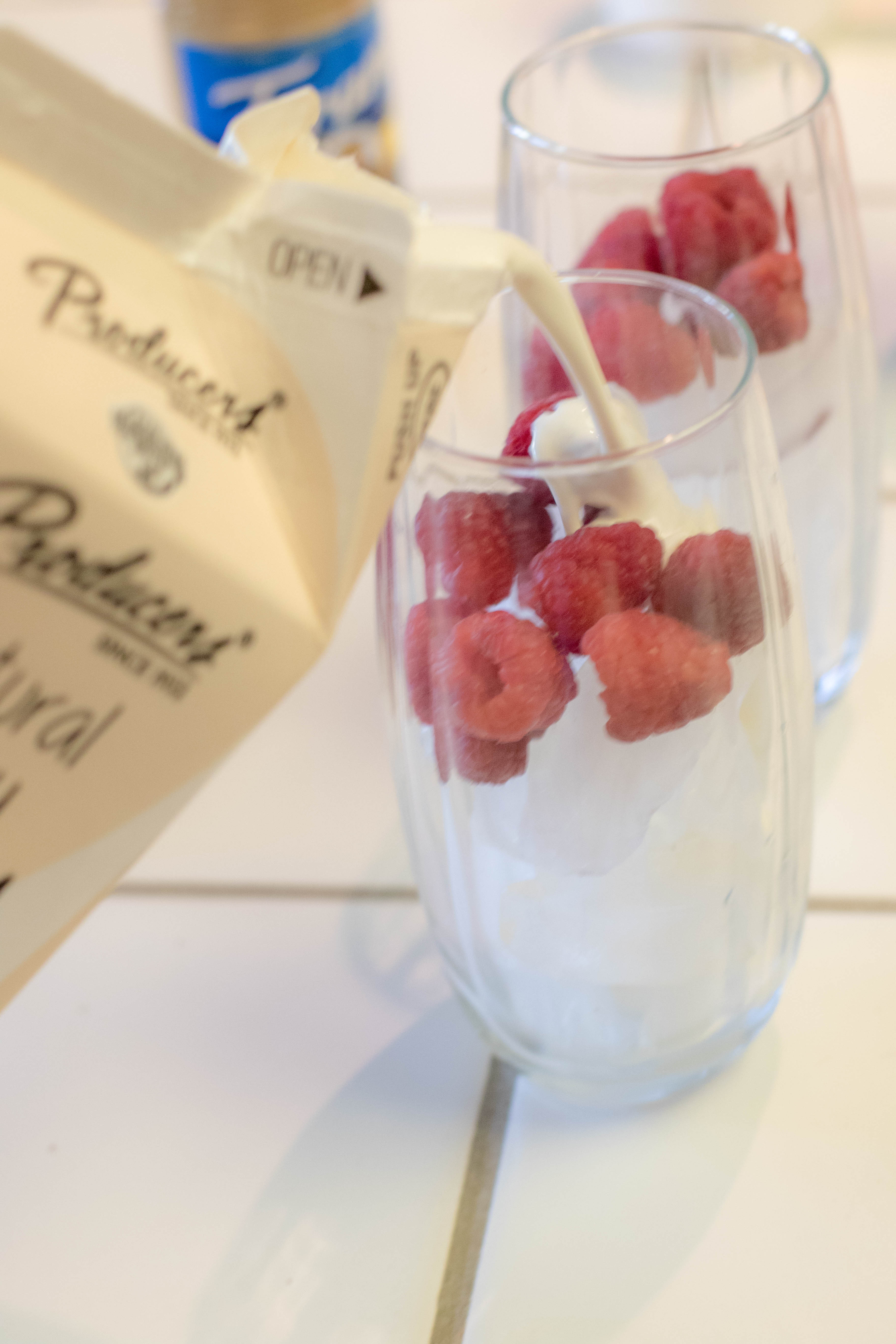 Natural Cream being poured into a clear glass with ice and raspberries on a kitchen counter.