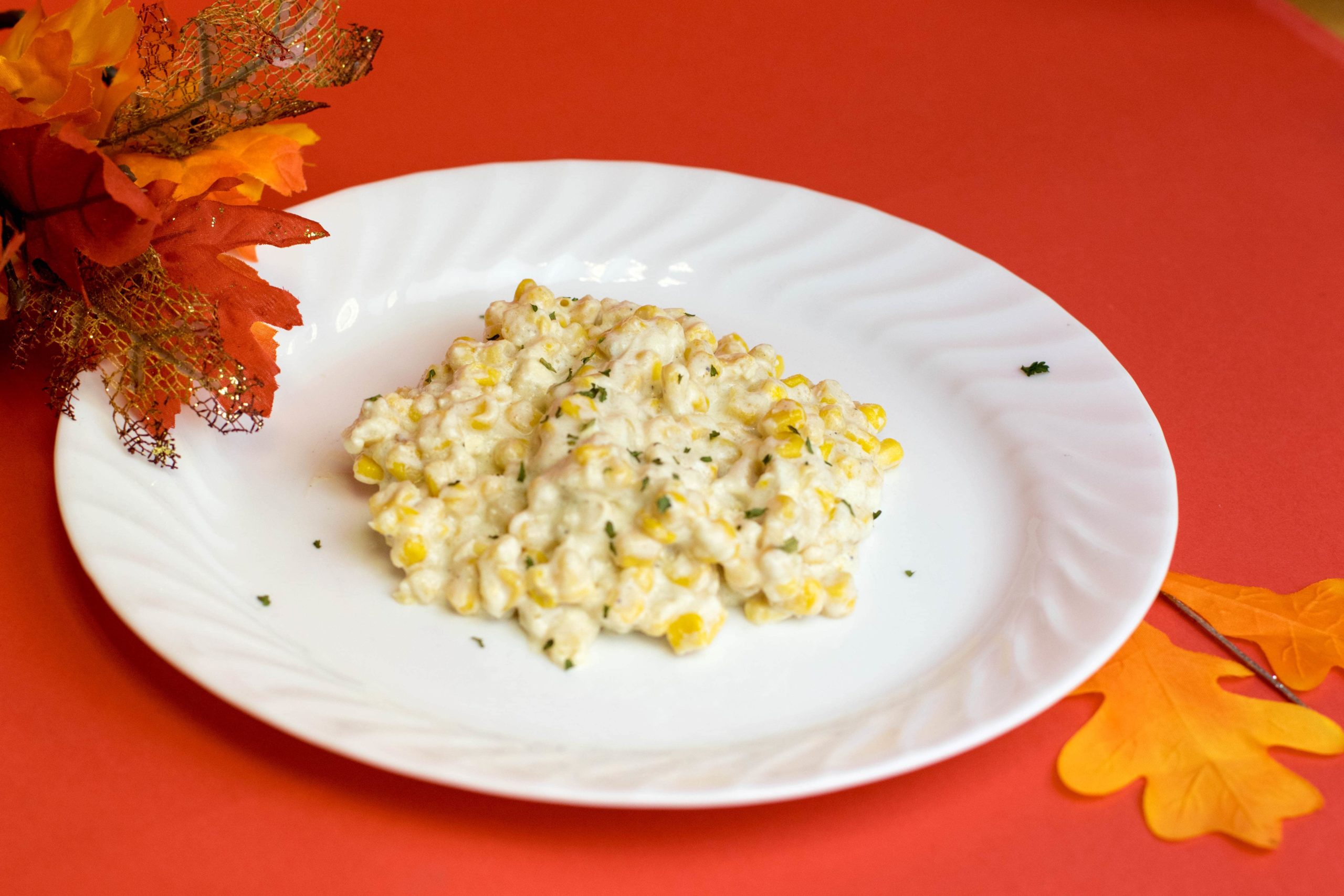 Creamed corn on a white plate laying on a flat red background surrounded by fall leaves.