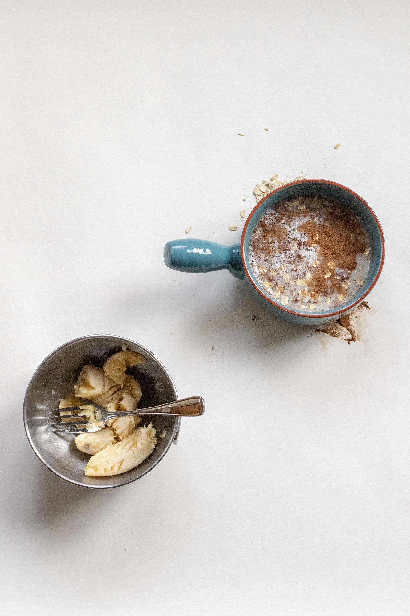 Mashed band with a fork in a metal bowl and egg whites with cinnamon and milk in another blue bowl. Both on a white background.