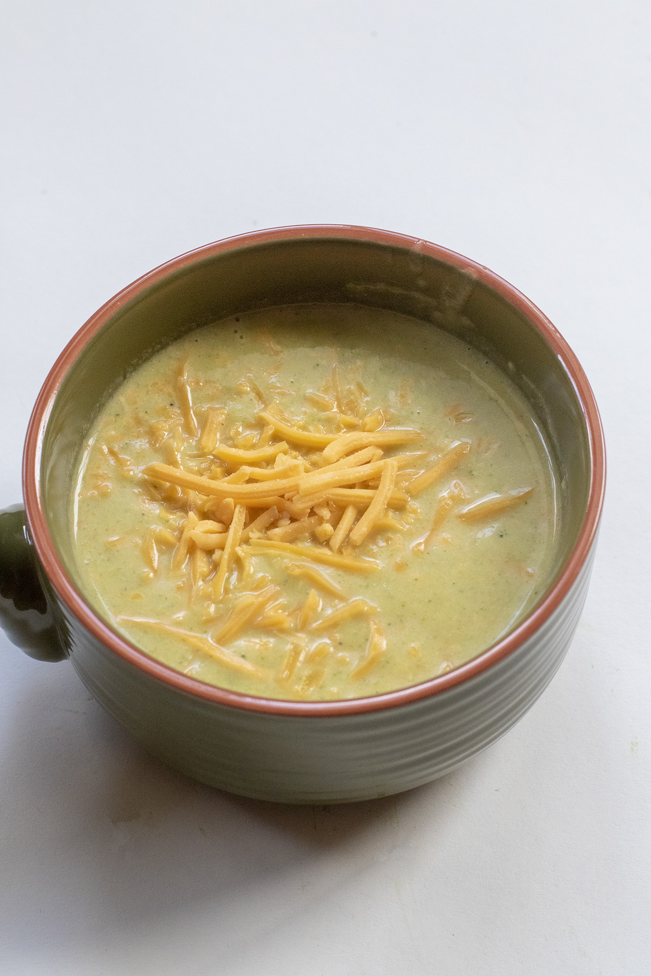 Broccoli Soup in a green bowl topped with shredded cheddar cheese laying on a flat white background.